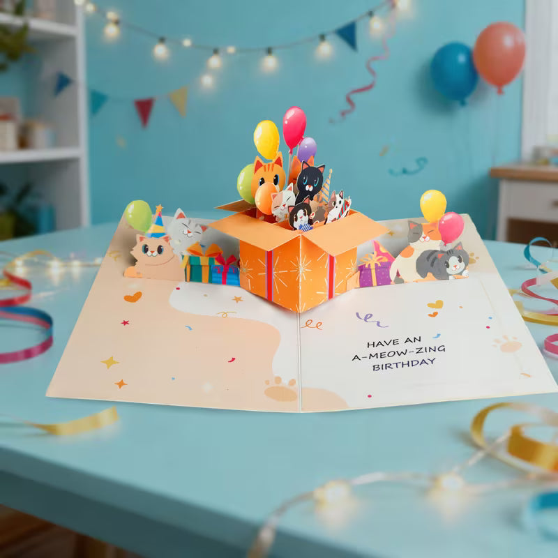 Avattu 3D pop-up -syntymäpäiväkortti, jossa oranssista lahjalaatikosta kurkistaa useita erivärisiä kissoja ja ilmapalloja sekä teksti Have an A-meow-zing birthday.