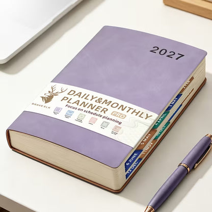 Tuotekuva lilasta A5-kokoisesta Daily & Monthly Planner Pro 2027 -kalenterista, jossa on pehmeä tekonahkakansi, värikkäät kuukausi-indeksit ja lila kirjoituskynä.   