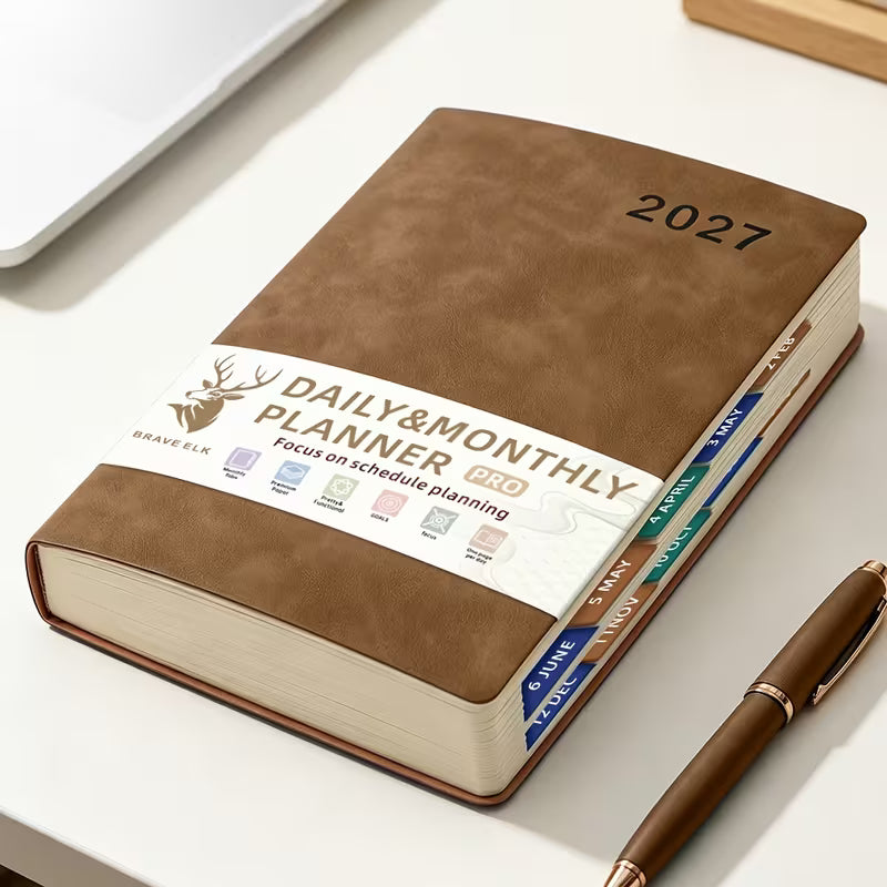 Tuotekuva lilasta A5-kokoisesta Daily & Monthly Planner Pro 2027 -kalenterista, jossa on pehmeä tekonahkakansi, värikkäät kuukausi-indeksit ja lila kirjoituskynä.   