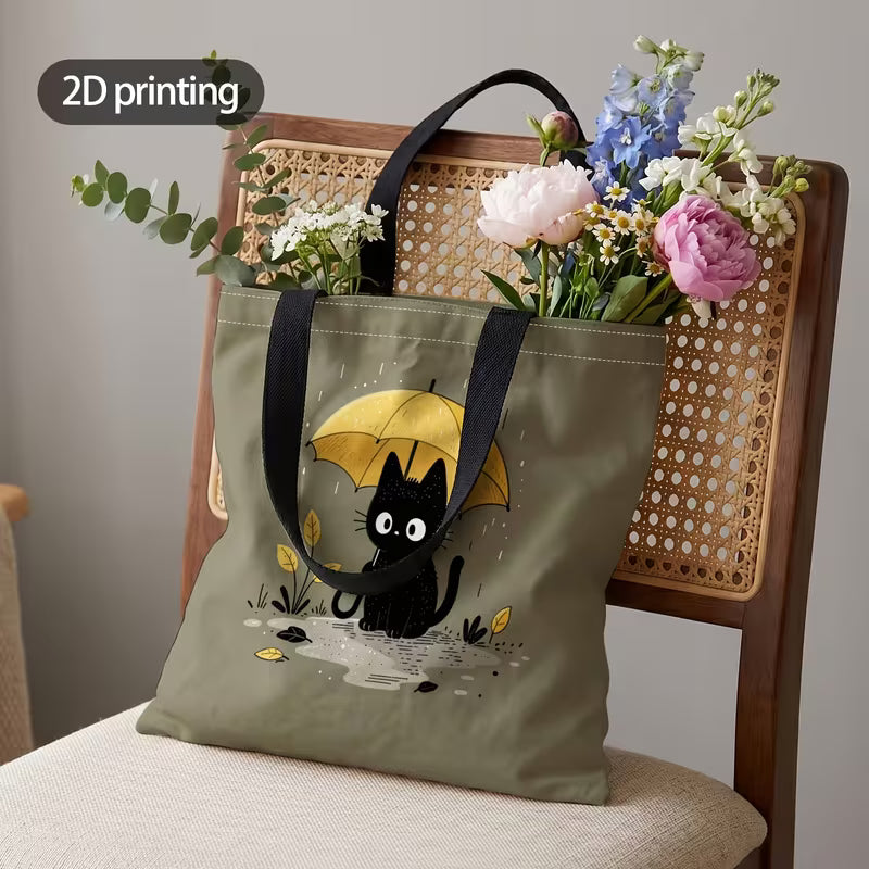 Tuotekuva salvianvihreästä kangaskassista, jossa on 2D-printtikuviointi mustasta kissasta keltaisen sateenvarjon alla sadepäivänä.