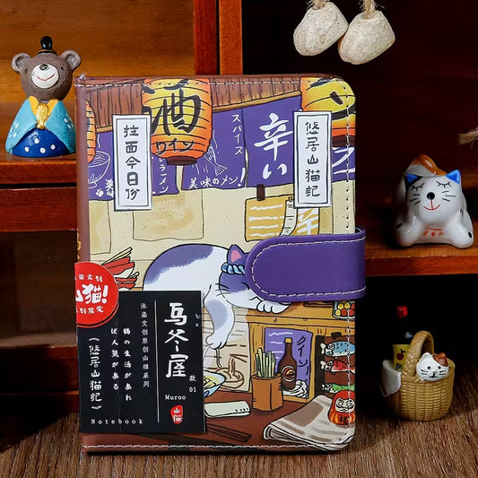 Japanilaistyylinen muistikirja, jonka kannessa on kissa perinteisessä kimonossa. Kirjassa on kultainen magneettisolki ja se on asetettu kirsikankukkien viereen.