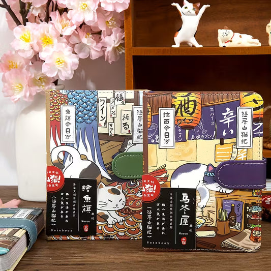 Japanilaistyylinen muistikirja, jonka kannessa on kissa perinteisessä kimonossa. Kirjassa on kultainen magneettisolki ja se on asetettu kirsikankukkien viereen.
