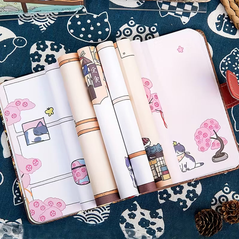 Japanilaistyylinen muistikirja, jonka kannessa on kissa perinteisessä kimonossa. Kirjassa on kultainen magneettisolki ja se on asetettu kirsikankukkien viereen.