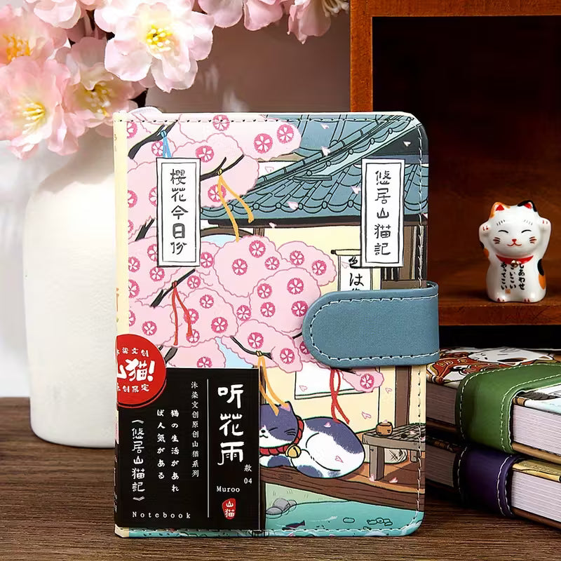 Japanilaistyylinen muistikirja, jonka kannessa on kissa perinteisessä kimonossa. Kirjassa on kultainen magneettisolki ja se on asetettu kirsikankukkien viereen.