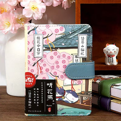 Japanilaistyylinen muistikirja, jonka kannessa on kissa perinteisessä kimonossa. Kirjassa on kultainen magneettisolki ja se on asetettu kirsikankukkien viereen.