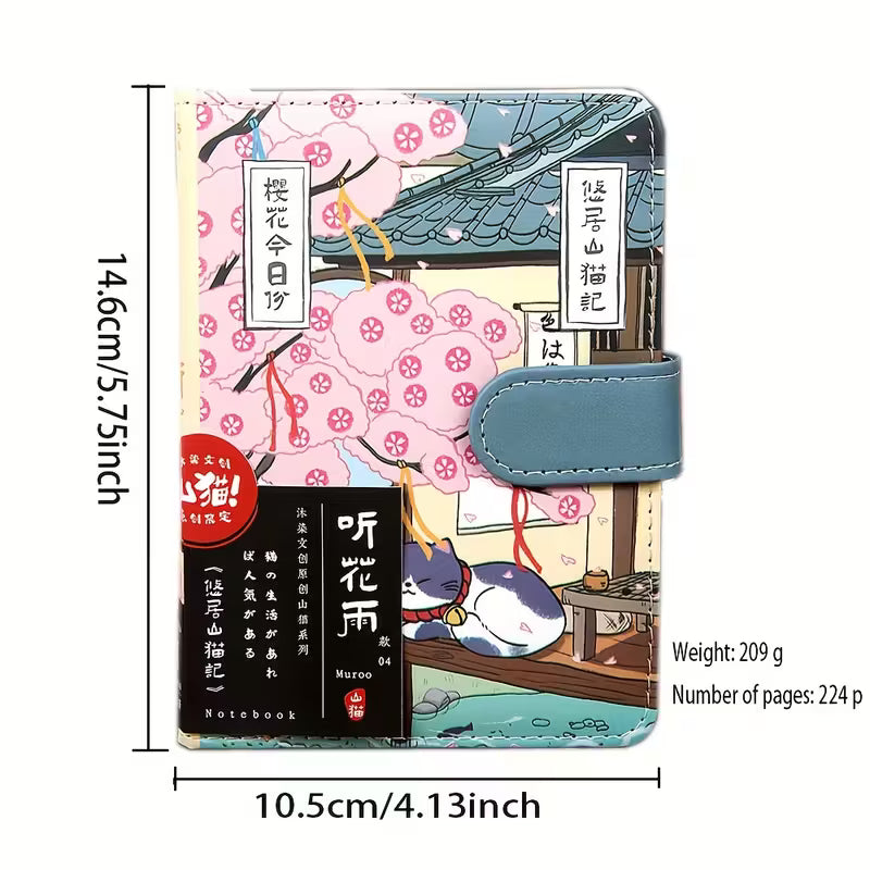Japanilaistyylinen muistikirja, jonka kannessa on kissa perinteisessä kimonossa. Kirjassa on kultainen magneettisolki ja se on asetettu kirsikankukkien viereen.