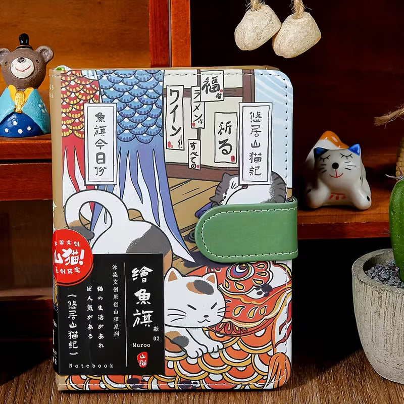 Japanilaistyylinen muistikirja, jonka kannessa on kissa perinteisessä kimonossa. Kirjassa on kultainen magneettisolki ja se on asetettu kirsikankukkien viereen.