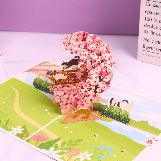 Avattu 3D pop-up -kortti, jossa on suuri vaaleanpunainen kirsikankukkakaari, leikkisä mustavalkoinen kissa ja oranssi kissa puutarhamaisemassa