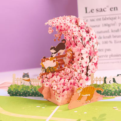 Avattu 3D pop-up -kortti, jossa on suuri vaaleanpunainen kirsikankukkakaari, leikkisä mustavalkoinen kissa ja oranssi kissa puutarhamaisemassa
