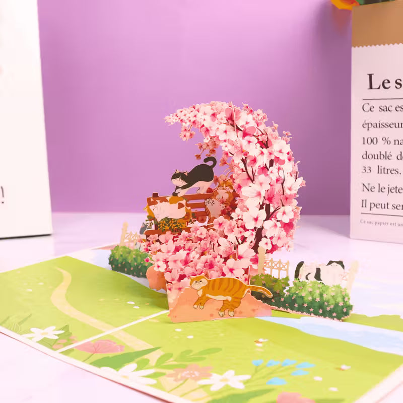 Avattu 3D pop-up -kortti, jossa on suuri vaaleanpunainen kirsikankukkakaari, leikkisä mustavalkoinen kissa ja oranssi kissa puutarhamaisemassa