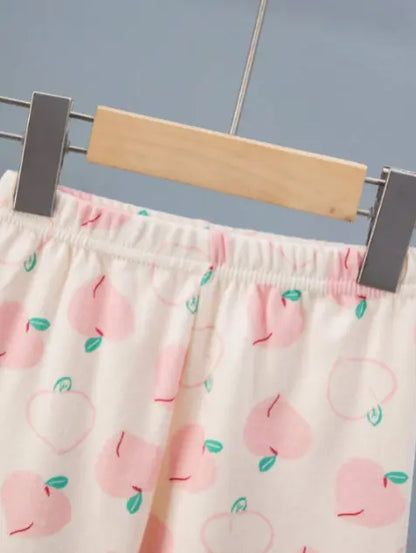 Vaaleanpunaisella persikkakuviolla koristeltu lasten puuvillapyjama, jossa on siro pitsireunus pääntiessä.