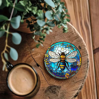 Yksittäinen pyöreä lasinalunen, jossa on koristeellinen mehiläiskuvio, aseteltuna puupöydälle teekupin alle.