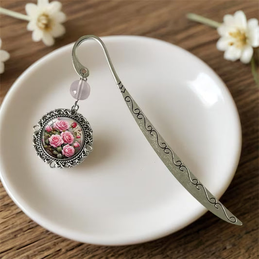 Hopeanvärinen metallinen kirjanmerkki, jossa on vaaleanpunainen helmi ja pyöreä mitali pinkeillä ruusuilla aseteltuna valkoiselle lautaselle.