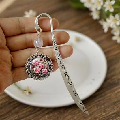 Hopeanvärinen metallinen kirjanmerkki, jossa on vaaleanpunainen helmi ja pyöreä mitali pinkeillä ruusuilla aseteltuna valkoiselle lautaselle.